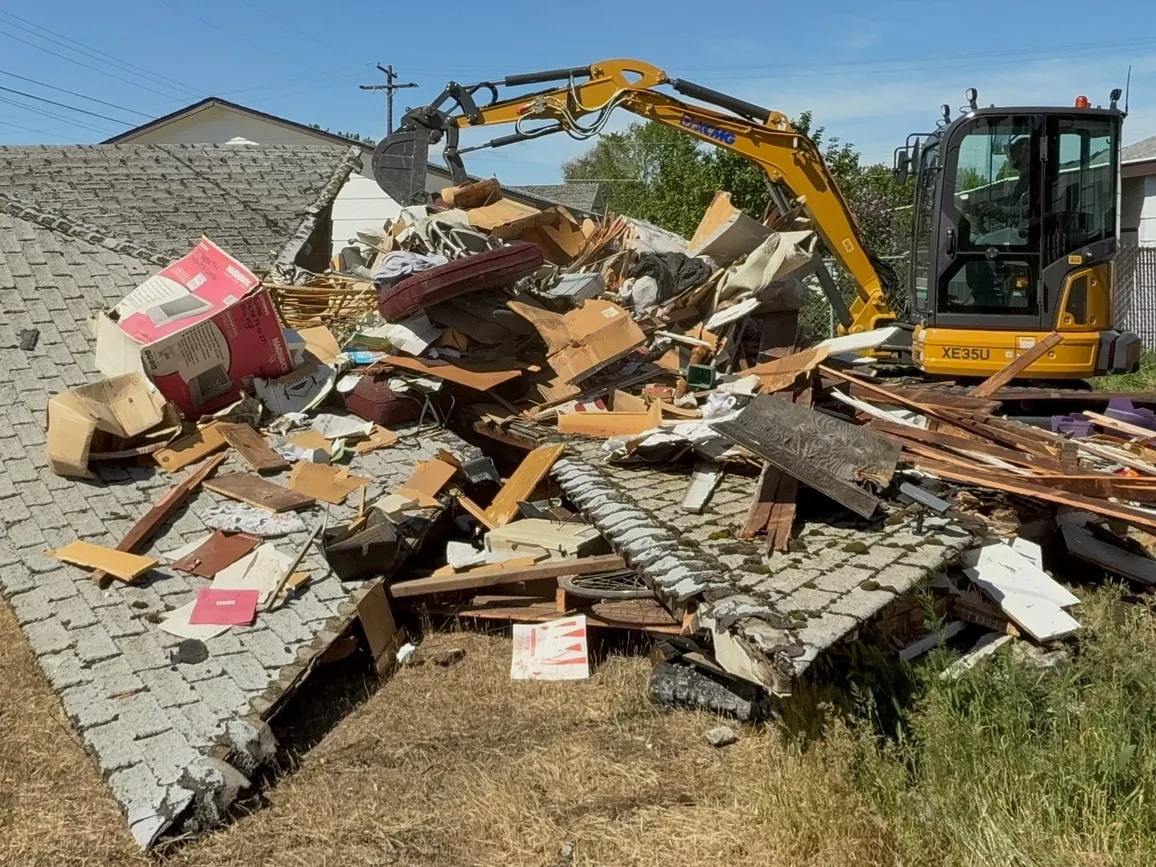 Mini excavator piling debris from a torn down detached garage, its shingled roof lying in the yard, building demolition