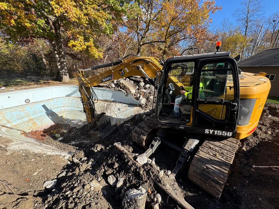 Excavator dropping broken concrete chunks into an old in ground pool shell, concrete removal