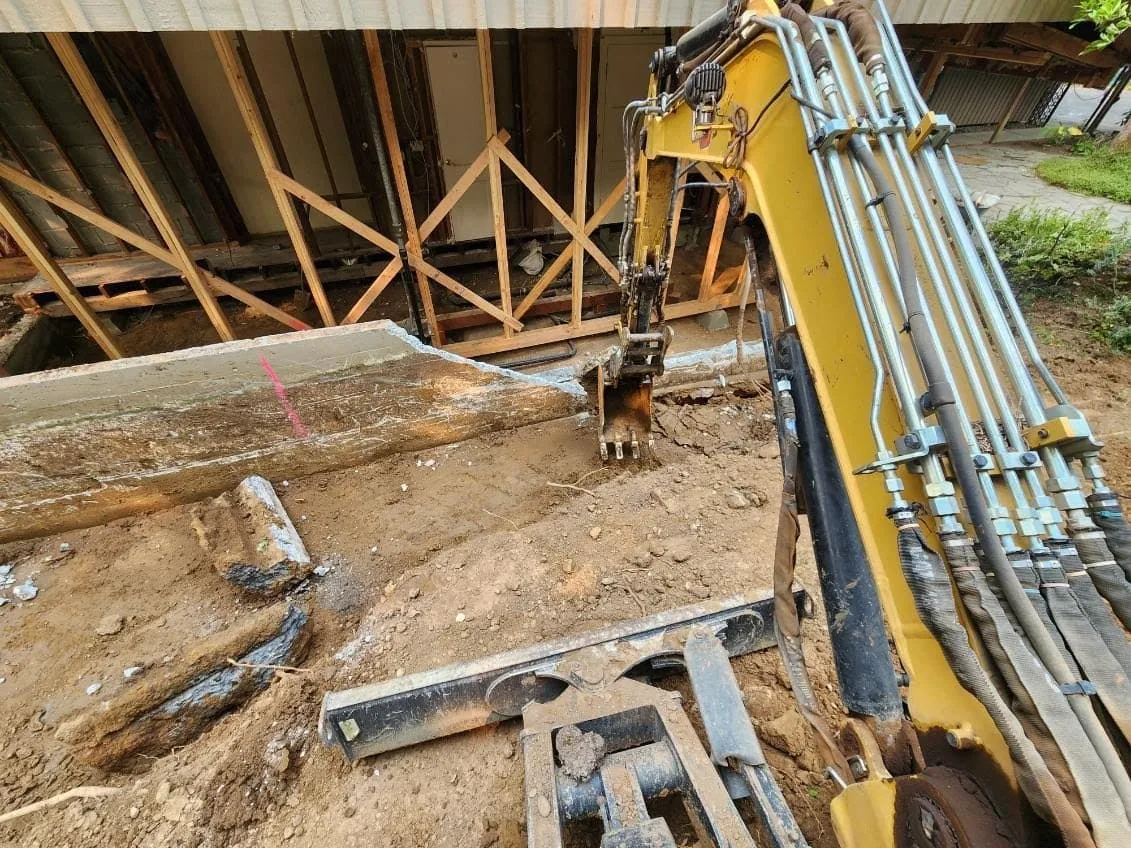 Mini excavator arm breaking out an old concrete footing under the raised floor framing of a house, concrete removal