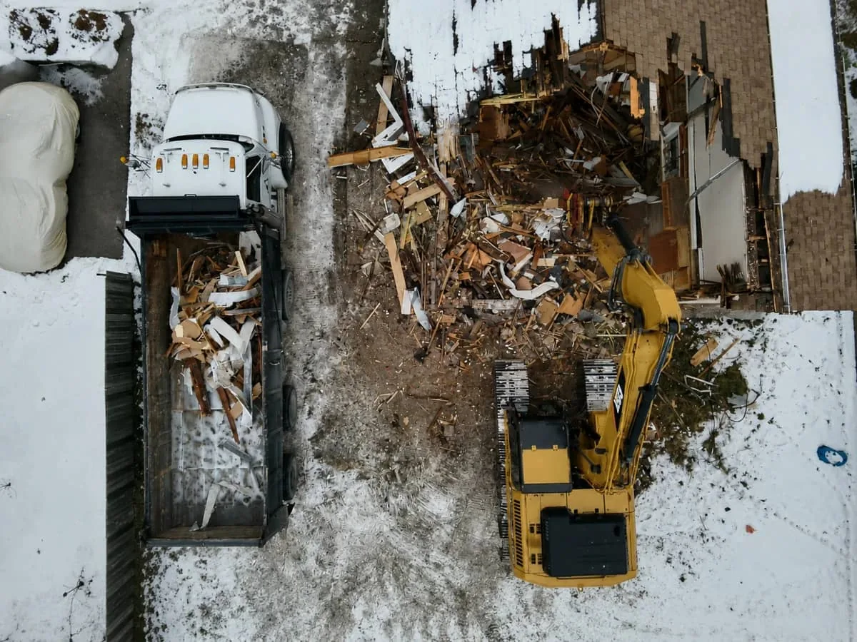 Aerial view of an excavator loading demolition debris into a dump truck beside a partly torn down house in the snow, debris removal