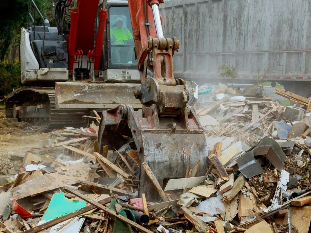 Excavator with a thumb sorting a pile of demolition debris beside a high sided dump trailer, debris removal