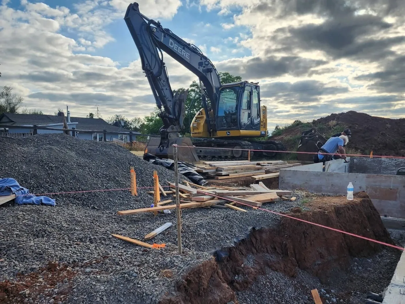Excavator at a house foundation dig in red clay with string lines and stacked lumber, excavation