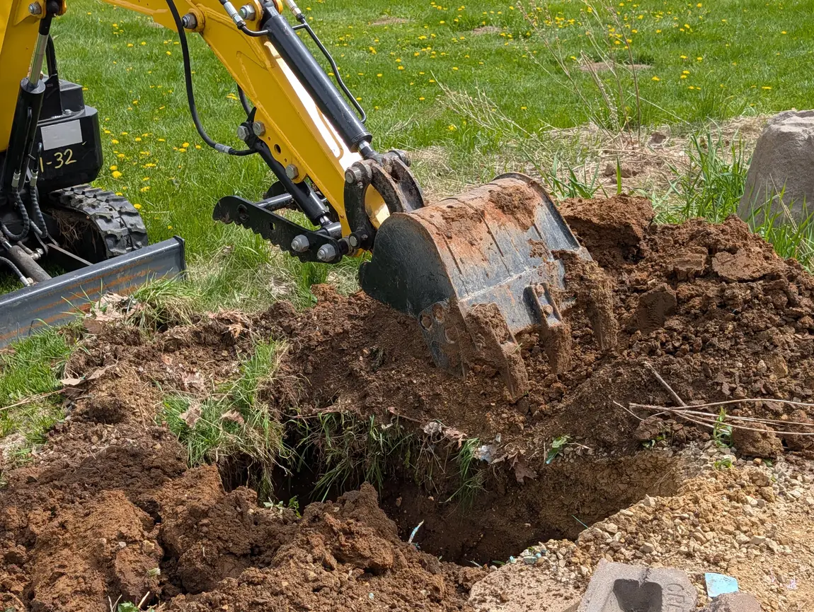 Small excavator digging a footing hole at the edge of a grassy yard, excavation