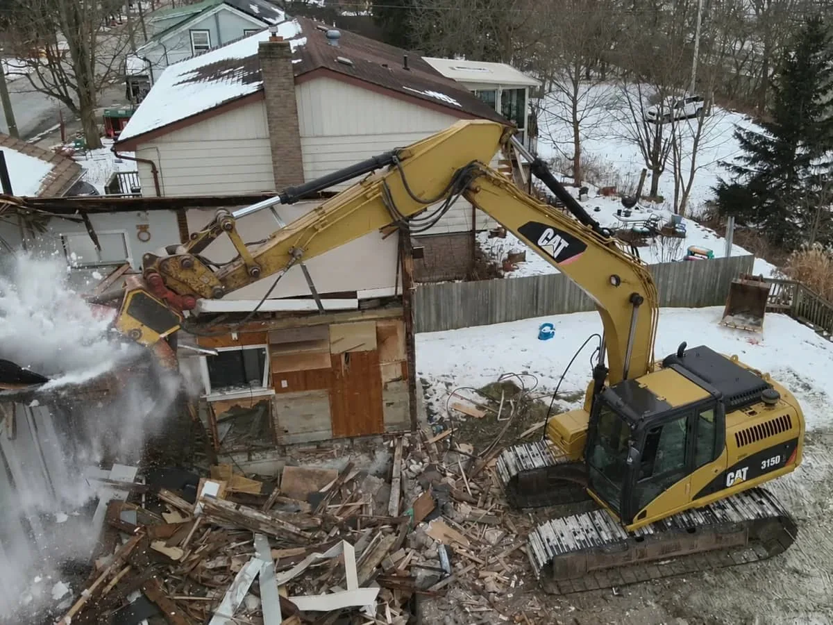 Excavator with a grapple tearing down a small house in winter snow, house demolition
