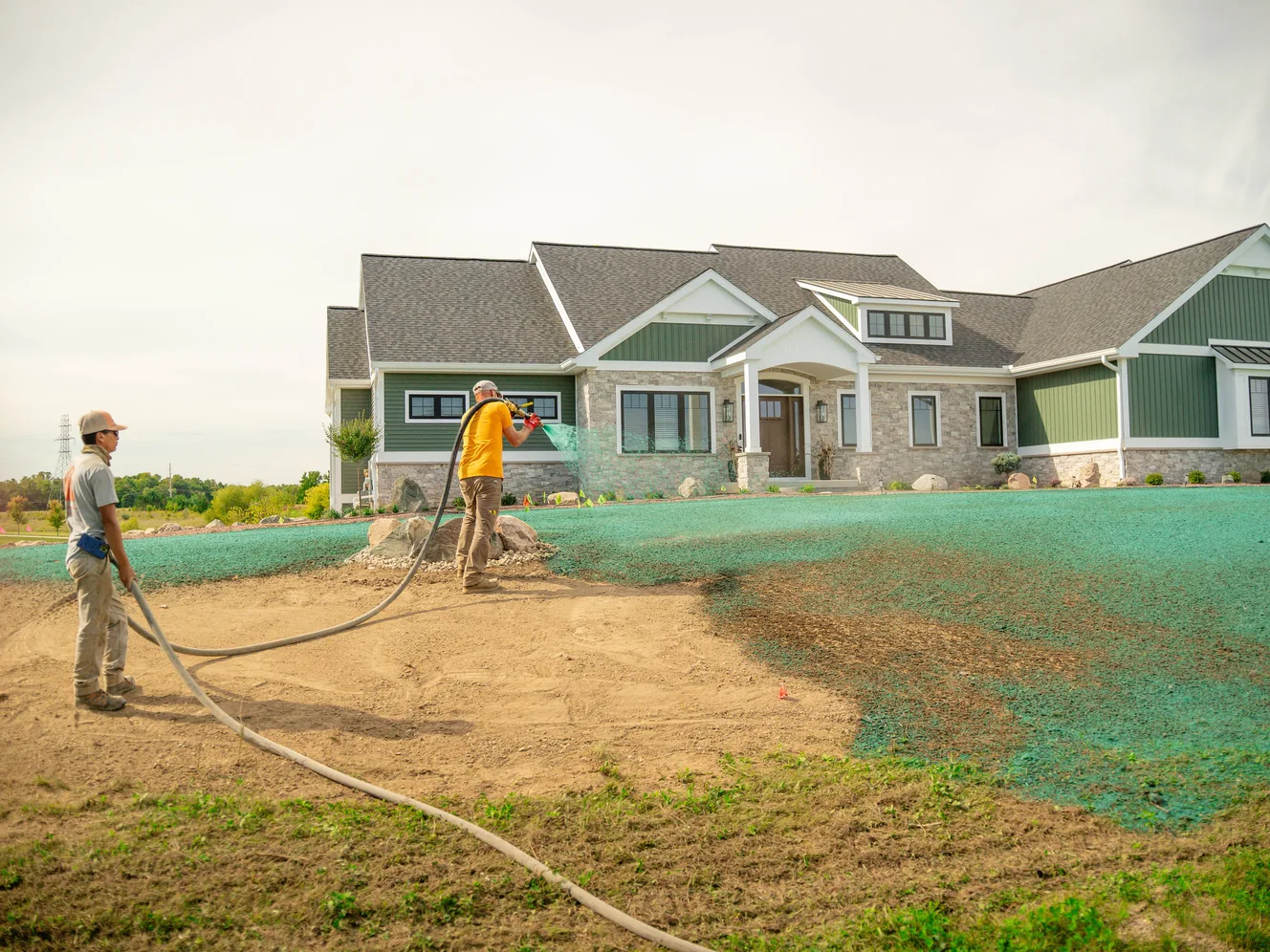 Two workers spraying green hydroseed mulch over the graded front yard of a new house, hydroseeding