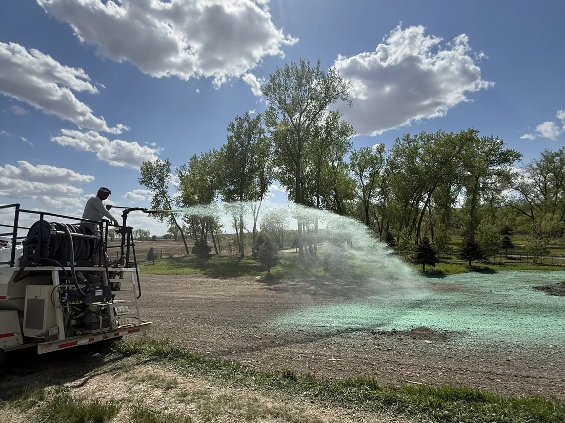 Worker on a hydroseeder truck spraying green slurry across a graded open lot, hydroseeding