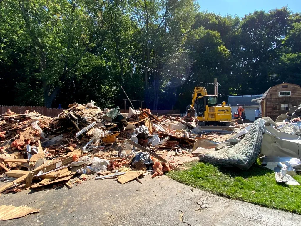 Debris pile of a torn down home with metal siding, insulation and a mattress, an excavator behind, mobile home demolition