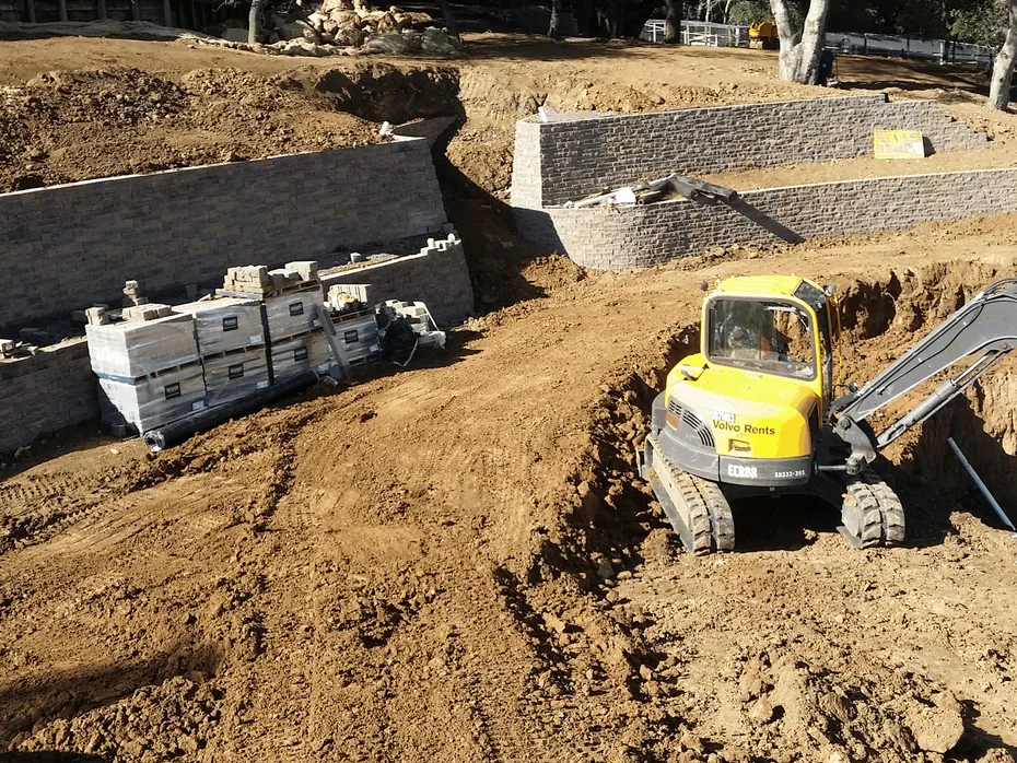Mini excavator digging below tiered block retaining walls under construction, retaining walls