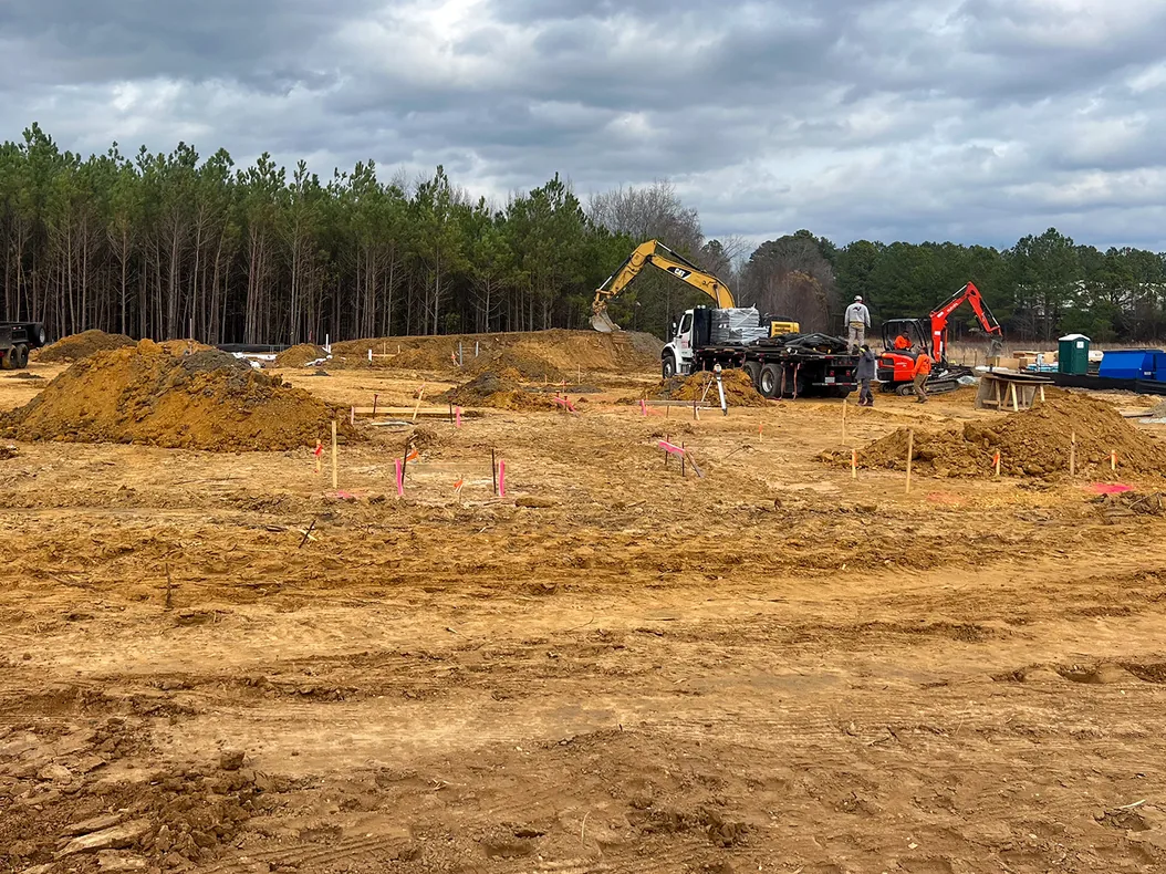 Rough graded building site with survey stakes and dirt piles, excavators in the distance, site development