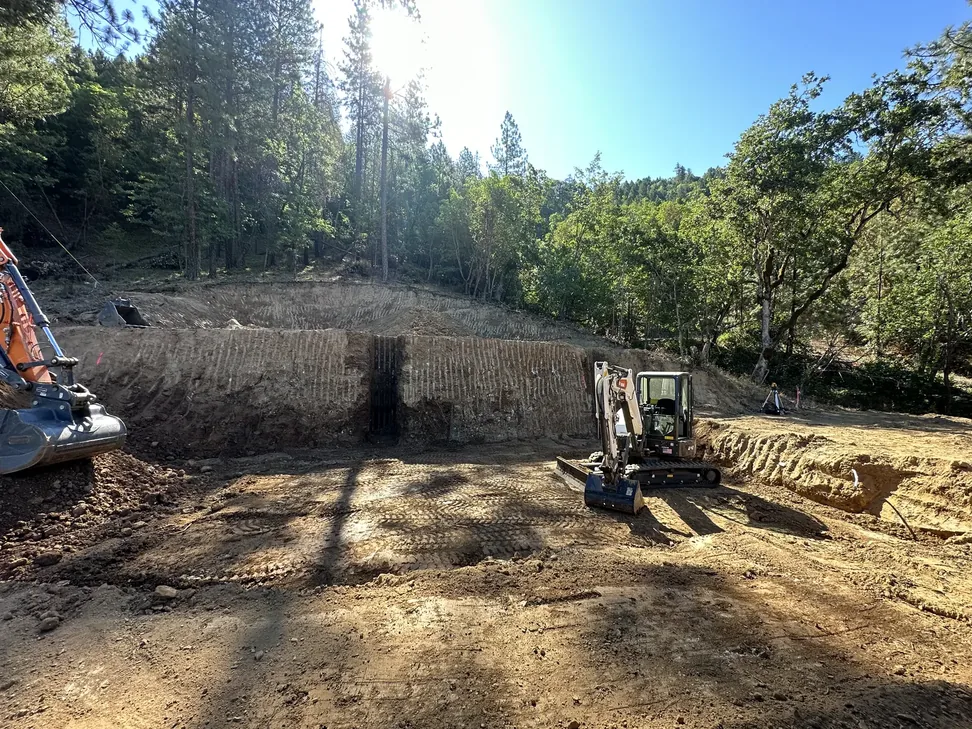 House pad cut into a wooded hillside with a small excavator on the pad, site development