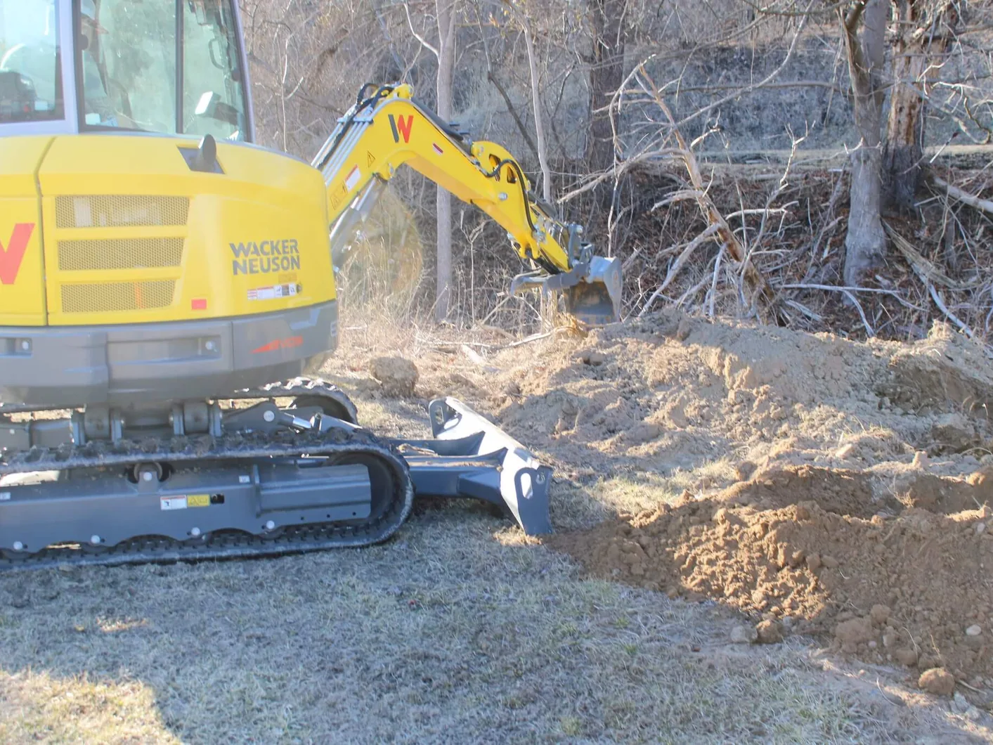 Mini excavator digging into a dirt bank beside bare winter brush, site development