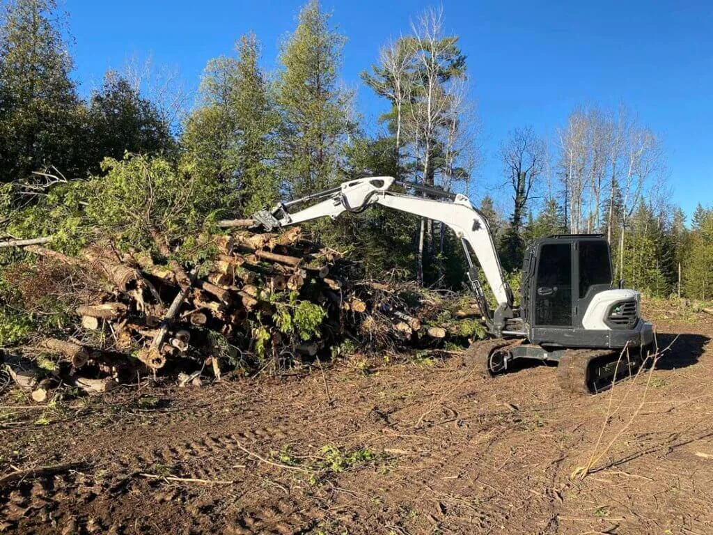Excavator with a thumb stacking cut tree trunks and brush on a cleared lot, land clearing